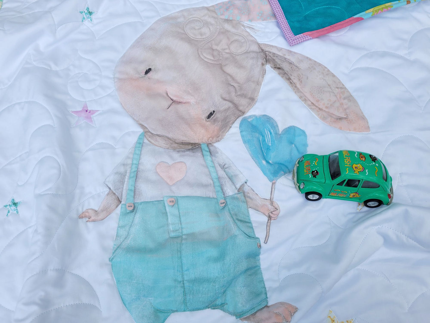 Cute baby quilt with bunny in overalls with a heart, blue balloon on white background with toy car sitting on it.