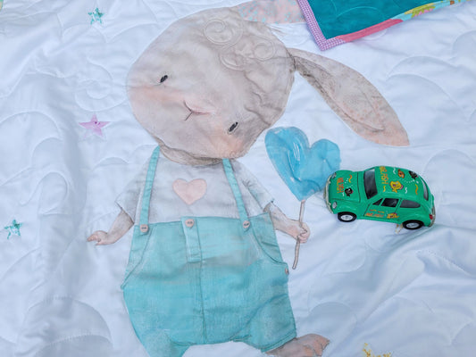 Cute baby quilt with bunny in overalls with a heart, blue balloon on white background with toy car sitting on it.
