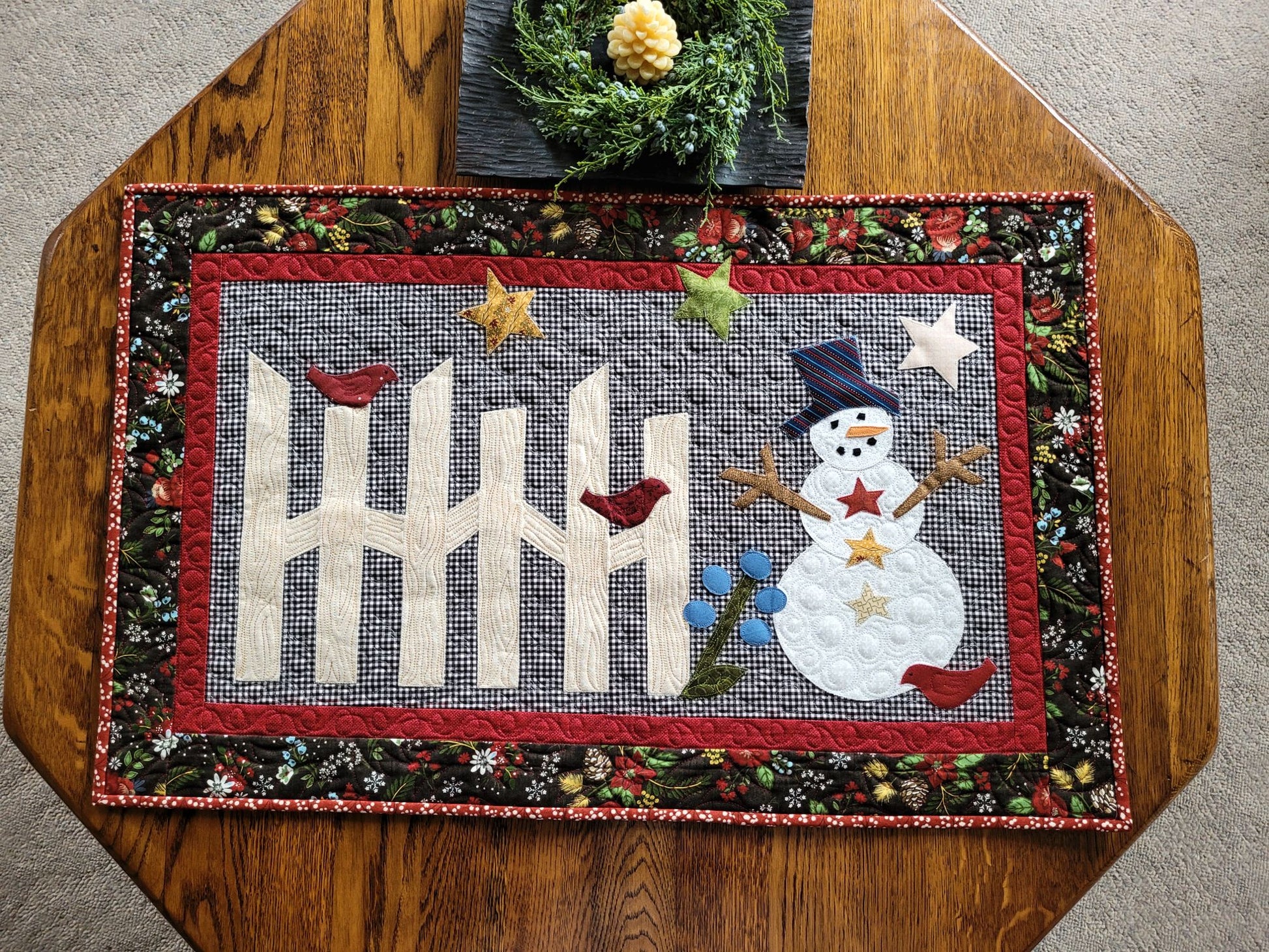 Cute snowman scene with rail fence, stars and birds on gingham background. The quilt has a small red border surrounded by black winter theme floral & pine cone print border. Quilts shown on hexagon wood table with candle.