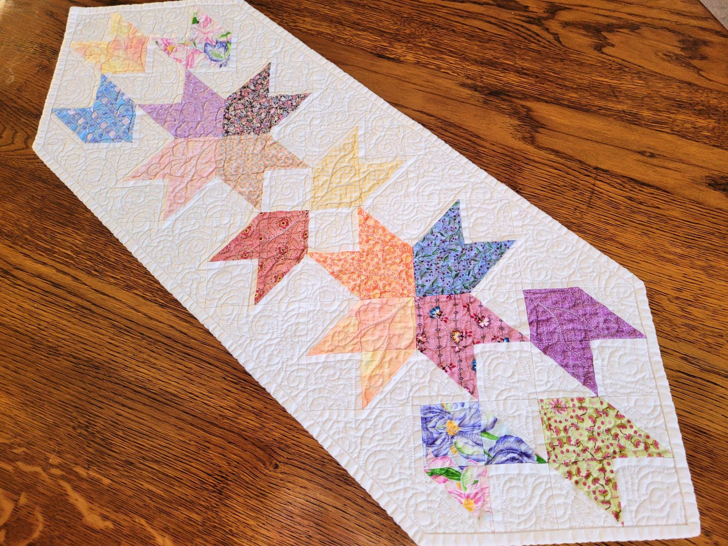 Quilted table runner with pastel colours on white background on wood surface.
