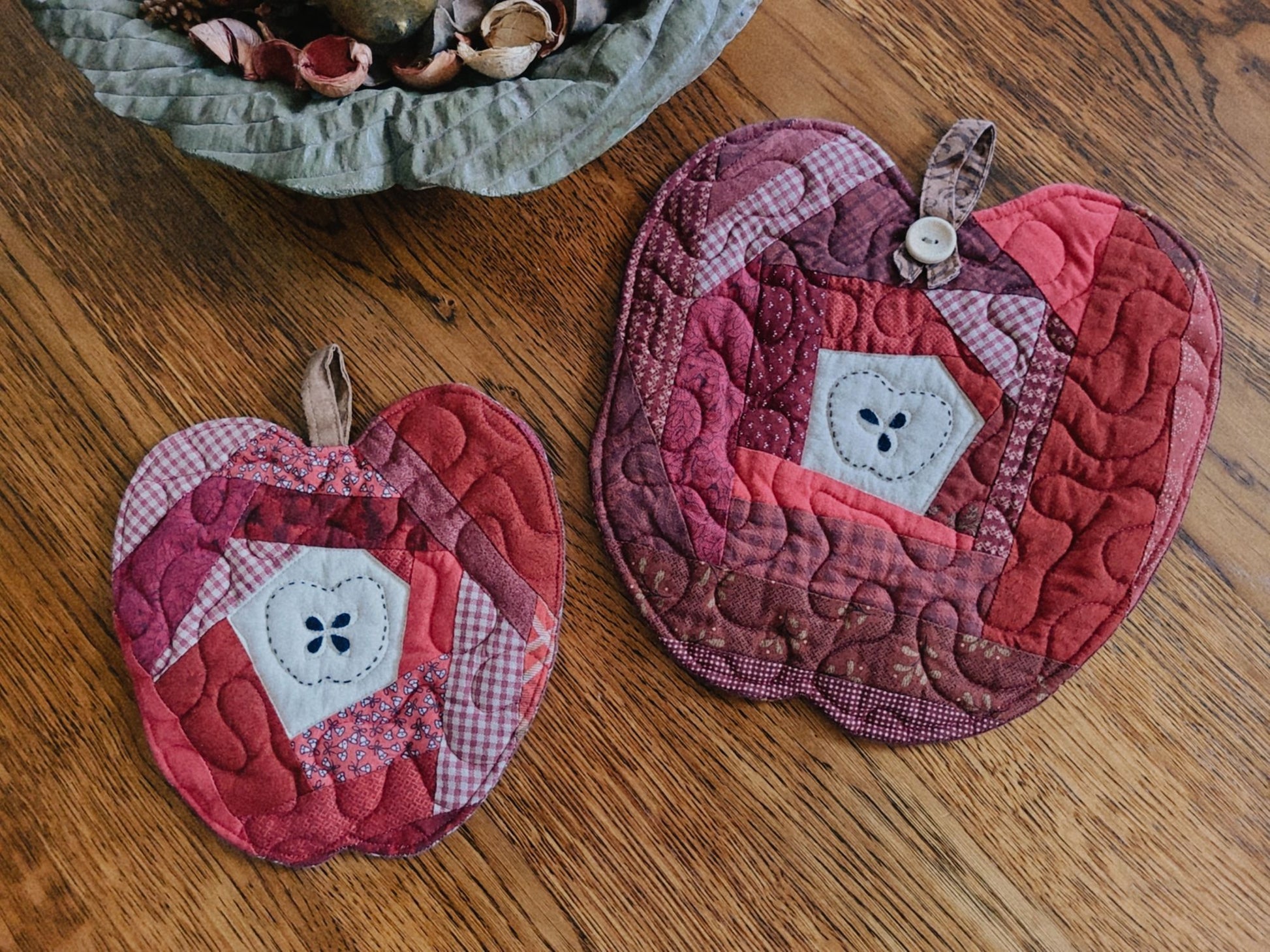 Two quilted fabric apple-shaped mats in shades of red and burgundy have a central white fabric with hand-stitched apple outline around applique seeds, displayed on a wooden tabletop.