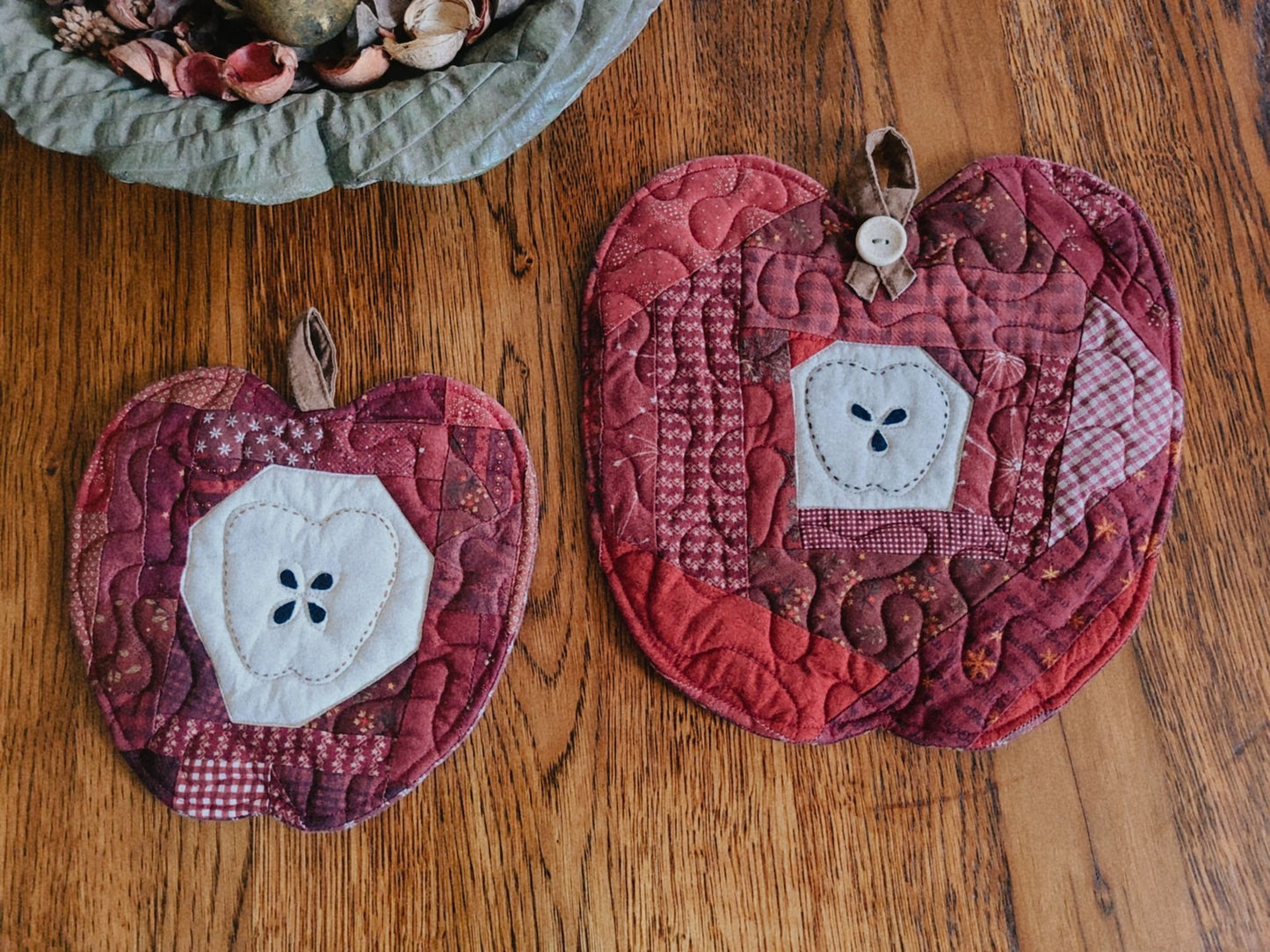 Two quilted apple-shaped quilts in shades of red, burgundy, and brown with white fabric apple centers, displayed on a wooden tabletop.