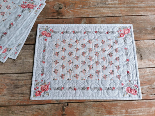 four quilted placemats in coordinating pale blue french floral fabrics.  Center block is a lattice style print with small sprigs of flowers evenly spaced throughout on a light blue background. The outer border has a trail of flowers along each side leading to the four cornerstones in a large scale floral print. Binding is a light blue and white pinstripe. 