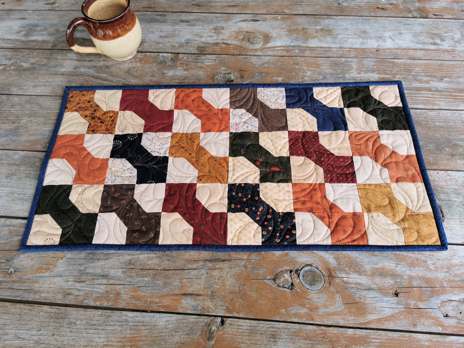 The quilted table runner is made of bowtie patchwork. Dark country florals are from collection called Paisley Park by Kansas Troubles. Bowties colours include country red, navy blue, brown, green, burnt orange, and antique gold. A scrappy beige background adds rustic charm. Dark blue binding is around the edge of the quilt.