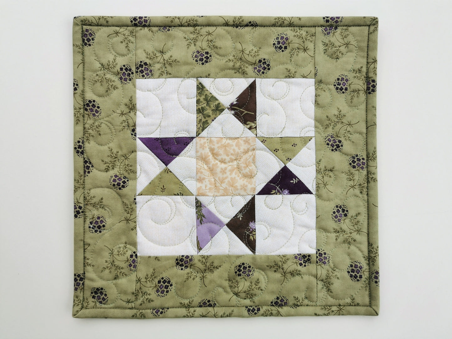 The scrappy star mini quilt shown on a plain white background. the outer border is a pretty spring green with small purple flowers. The interior star has a cream background with the star points made of scrappy fabrics that coordinate with the outer border.