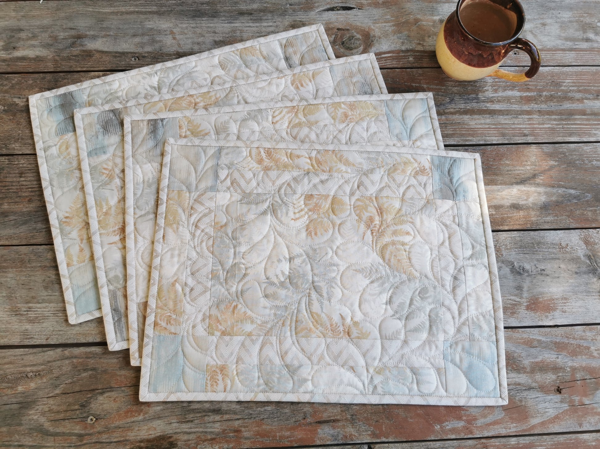 four quilted placemats in neutral beige and slate blue fabrics with a muted abstract fern pattern. 