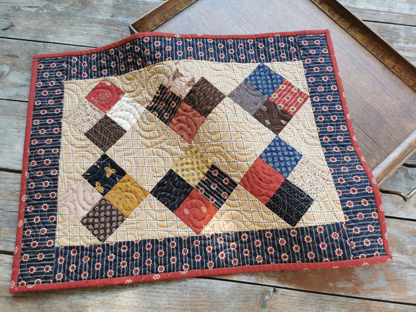 This small scrap quilt features civil war fabrics  in rustic country prints. Scrappy four patch blocks are in the center on a beige background. A navy blue border with small red flowers surrounds the center blocks. A thin red binding to finishes the quilt. Perfect to use as a table topper.