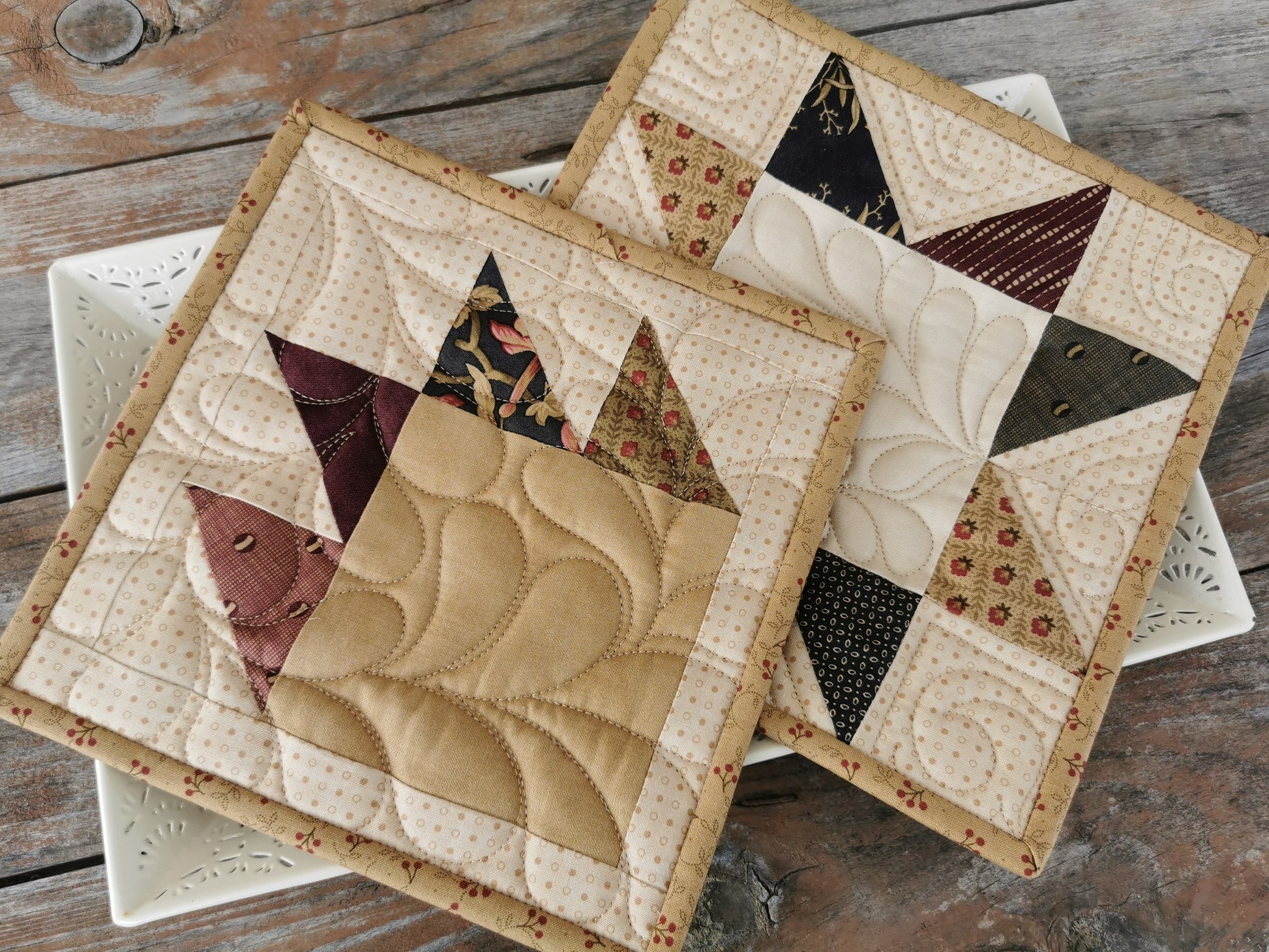 Set of potholders shown on a white tray. Top potholder has bear paw piecing and the one underneath is a patchwork star. Colors are  beige tones with country colors of magenta, coral red, charcoal, tan. 