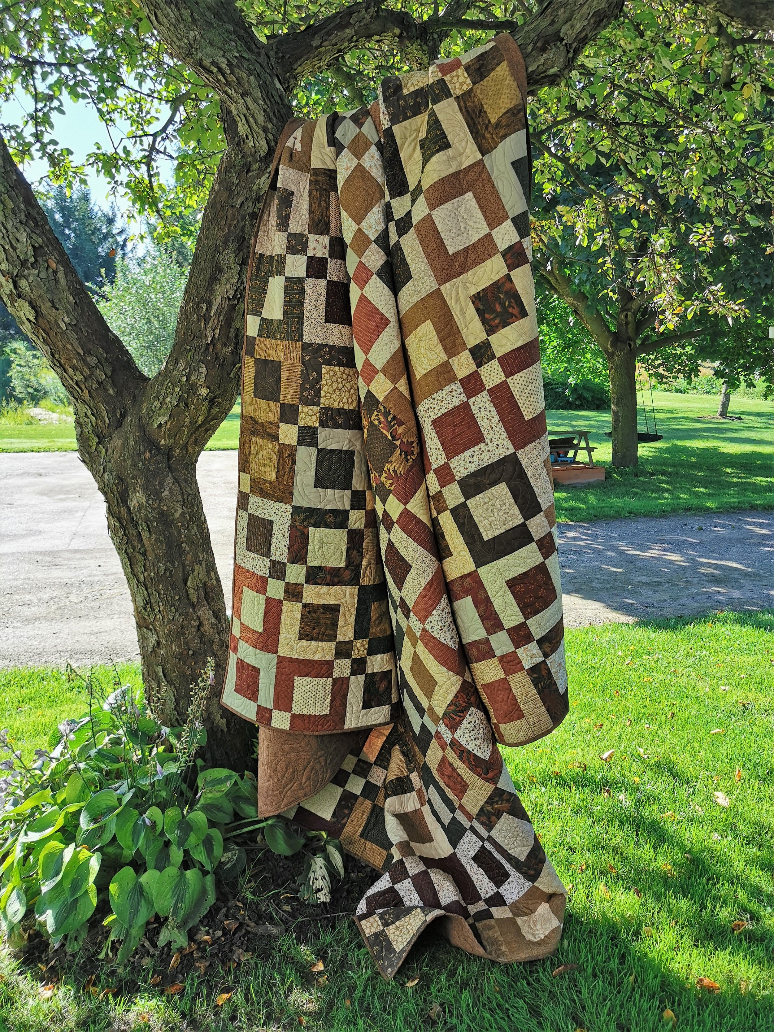 This patchwork queen bed quilt is shown hanging over a tree branch. The rustic colors include brown, green, beige, red, orange, gold. 