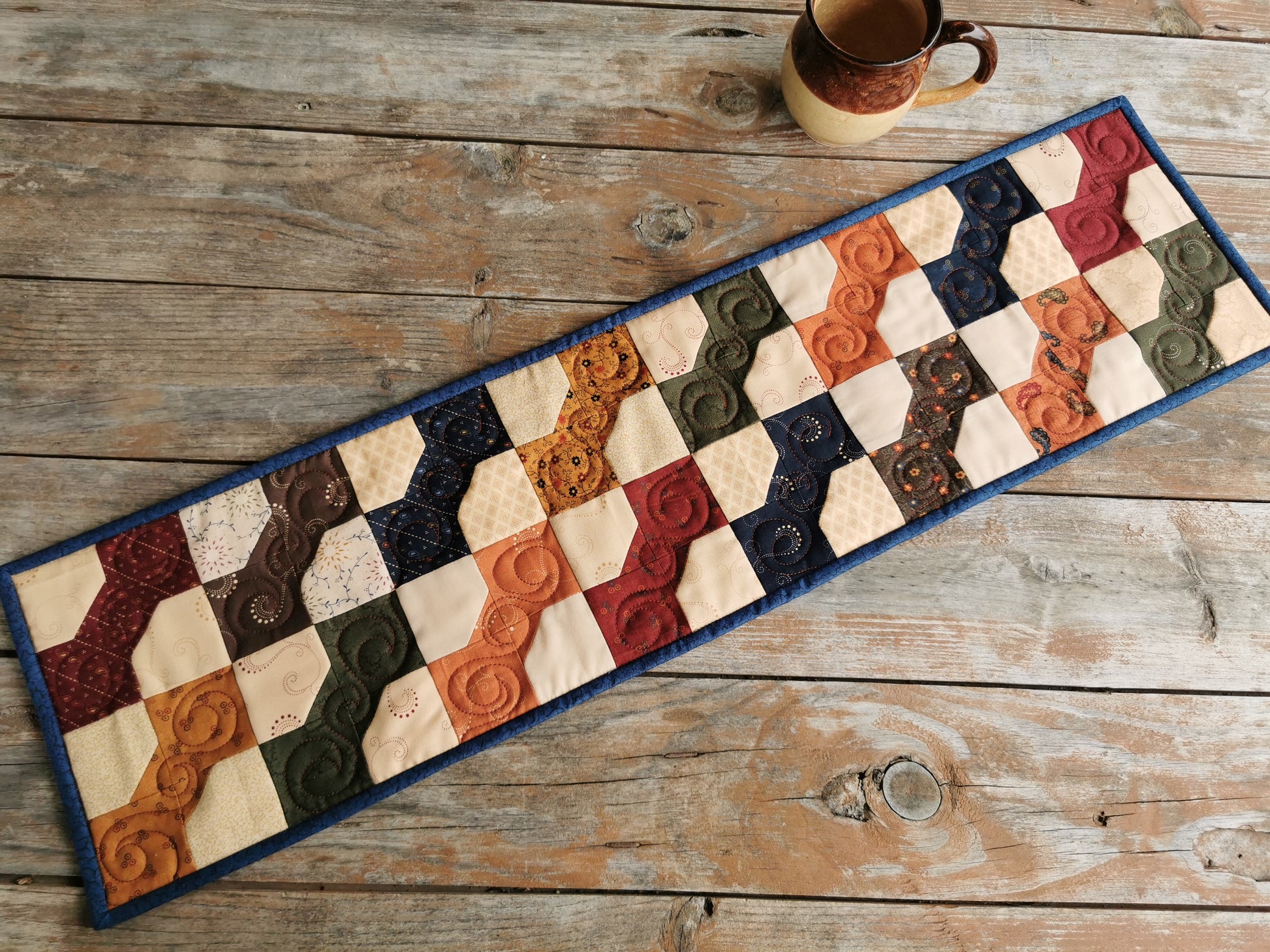 This narrow table runner with bowtie patchwork has  lovely dark country colours in a beige background. Each bowtie is a different fabric from a coordinating collection called Paisley Park by Kansas Troubles. The pretty country florals in the bowties include country red, navy blue, brown, green, burnt orange, antique gold. A dark blue binding is used around the edge.