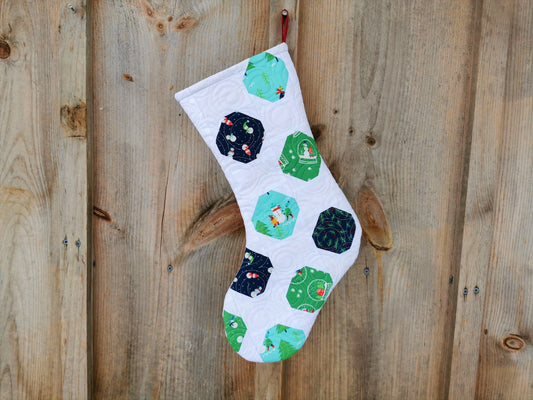 this heirloom quilted stocking features winter themed fabrics in navy, teal  and green colors on a white background. Fabrics have snowmen, pine trees, snow globe, pine branches with berries. 