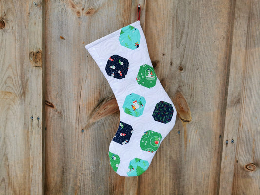this heirloom quilted stocking features winter themed fabrics in navy, teal  and green colors on a white background. Fabrics have snowmen, pine trees, snow globe, pine branches with berries.