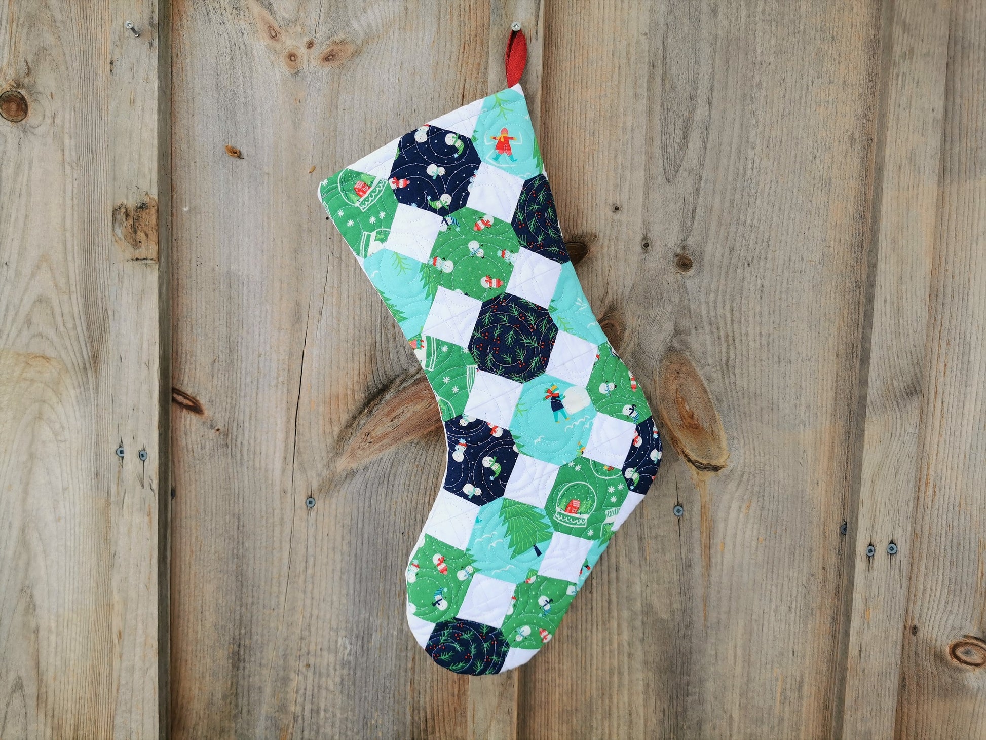 This quilted stocking is pieced with winter themed fabrics in navy, green, and light teal. The fabrics designs have snowmen, pine trees, snow globe, snowflakes, pine branches and berries. 