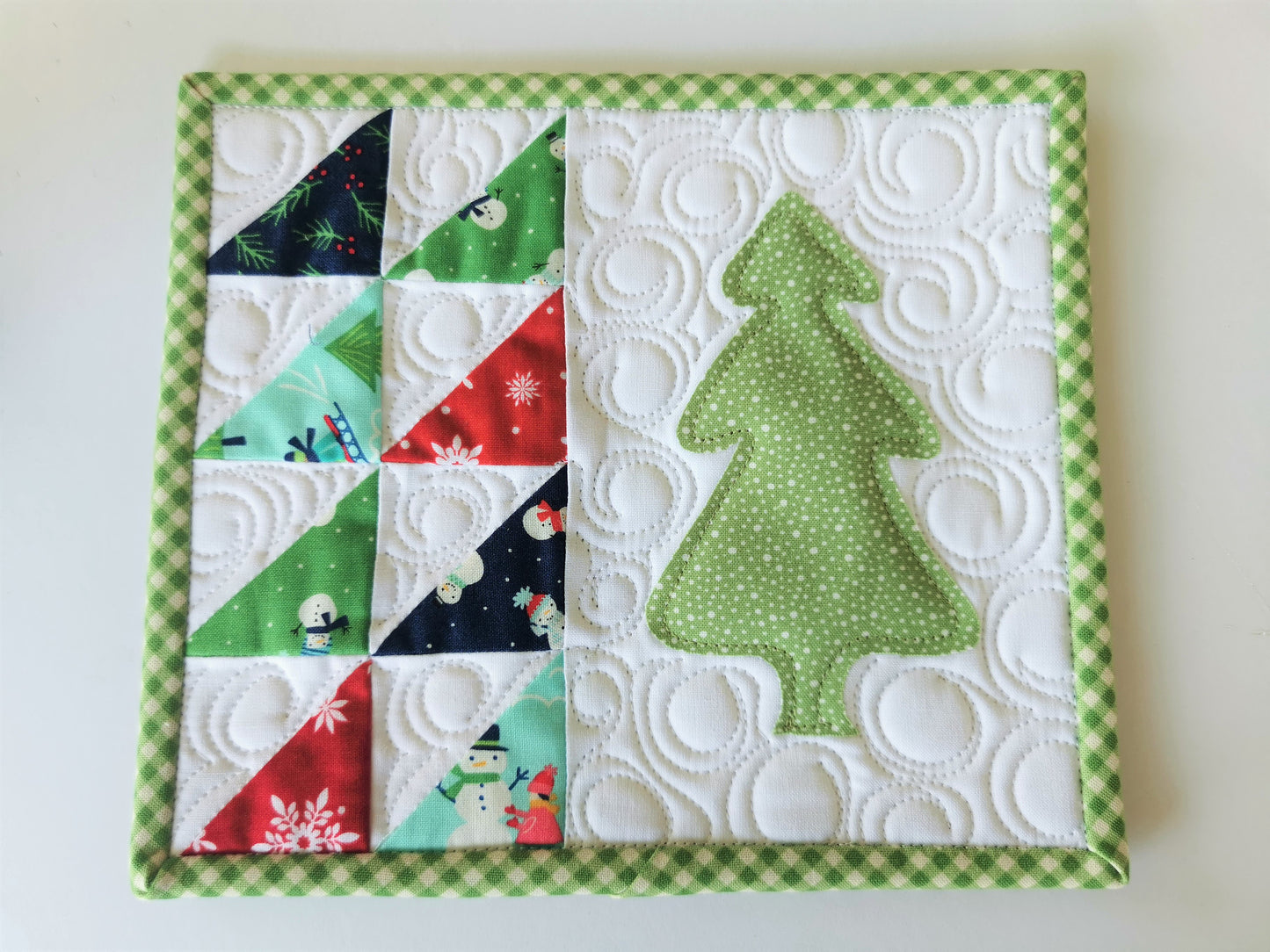 This mini christmas tree quilt features winter theme fabrics on white background. Small triangle patchwork down the left side with a green polka dot christmas tree on the right. Binding is a green gingham.