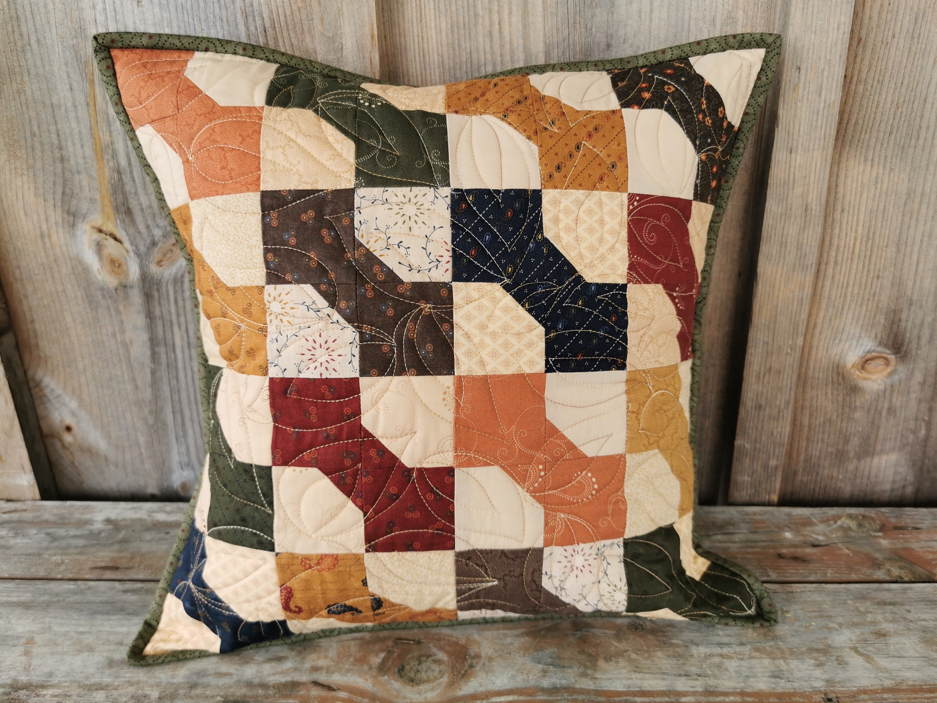 This quilted toss cushion has a country feel with the colorful bowtie patchwork. Bowties colors are:  navy, brown, burnt orange, forest green, antique gold and country red. The beige background has a scrappy look using various coordinating fabrics. A lovely, rustic country look. 