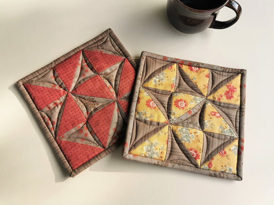 the quilted patchwork potholders have coordinating fabrics, one is pieced with tan and coral pink, and the other with tan and a golden yellow floral.