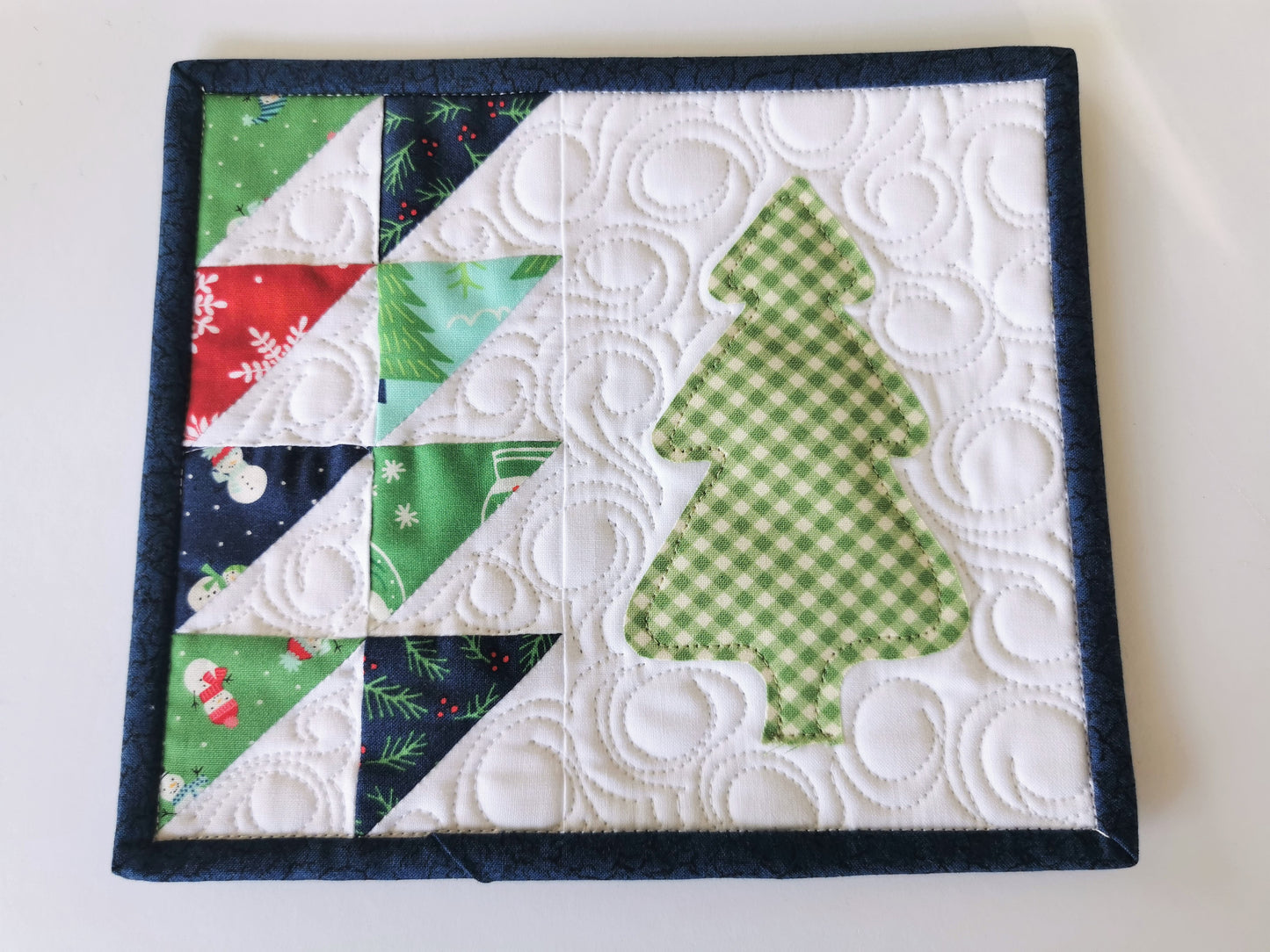 This mini christmas tree quilt features winter theme fabrics on white background. Small triangle patchwork down the left side  with a green gingham christmas tree on the right. Binding is a dark navy. 
