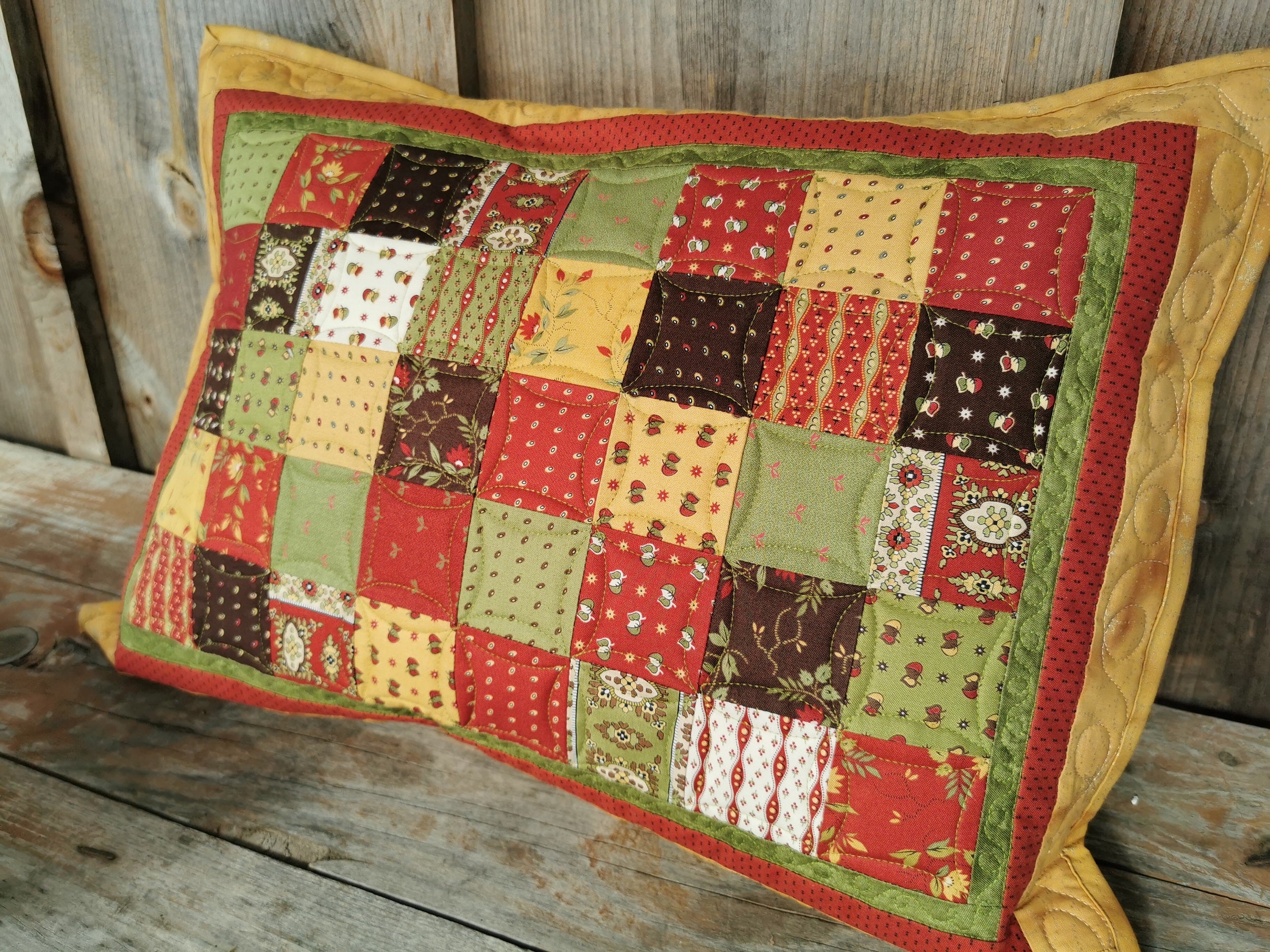This rectangle throw pillow is shown propped on a wooden table. Cushion colors are somewhat Christmassy. Simple patchwork squares are in the colors red, gold, green and a touch of dark brown and are warm and welcoming.