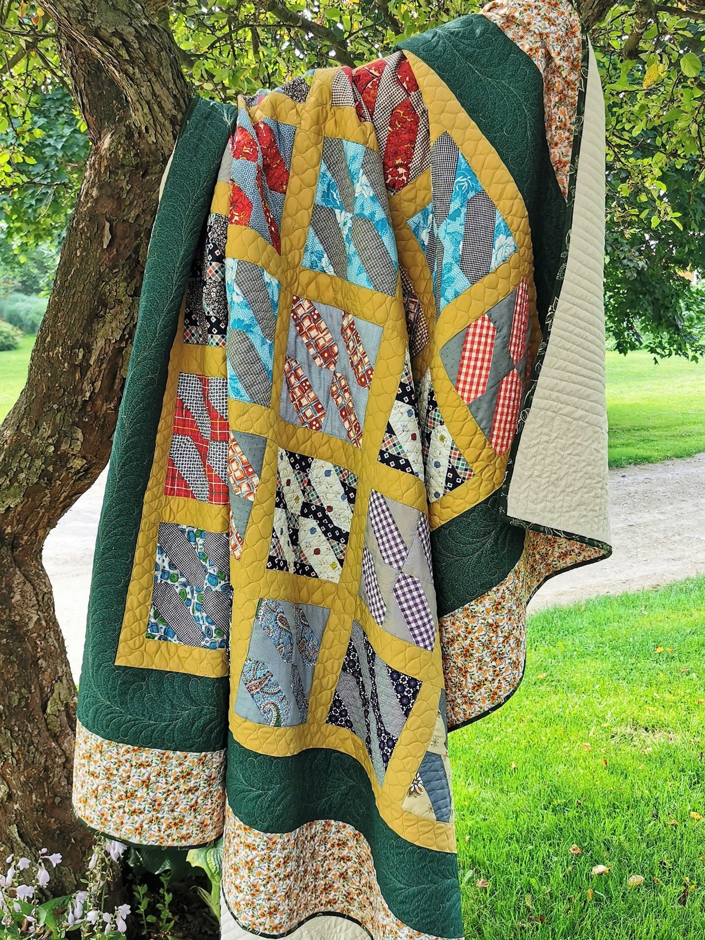 This large queen vintage bed quilt it shown in outdoor light, draped over the branch of a crab apple tree.  The middle of the quilt top has the vintage patchwork blocks set in a mustard yellow grid pattern. The interior grid is surrounded by a dark green border then a beige floral border around the outside