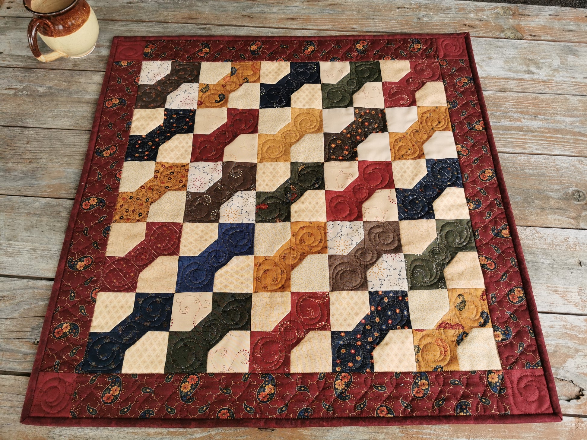 This square quilted table runner is made of bowtie patchwork surrounded by a deep country red floral print. Each bowtie is a different color from a collection of coordinating fabrics called Paisley Park by Kansas Troubles. This gives a lovely scrap quilt look. Bowtie colors include, deep blue, brown, red, burnt orange, green, antique gold.