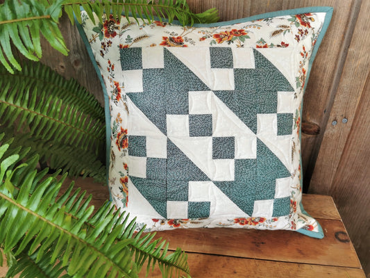 This quilted pillow is shown on a wooden table beside a fern, in outdoor natural light. Teal and white patchwork with a complimentary floral border is striking.