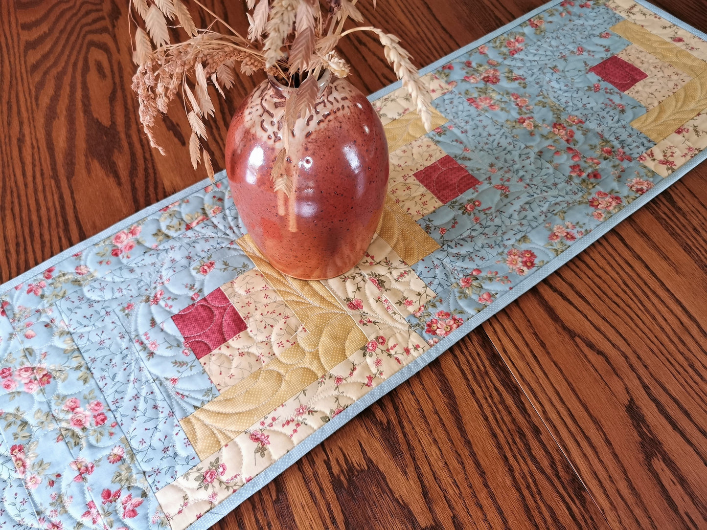 This quilted log cabin table runner features floral fabrics in soft colors. Coordinating fabrics are teal blues, golden yellows with a touch of coral pink.  