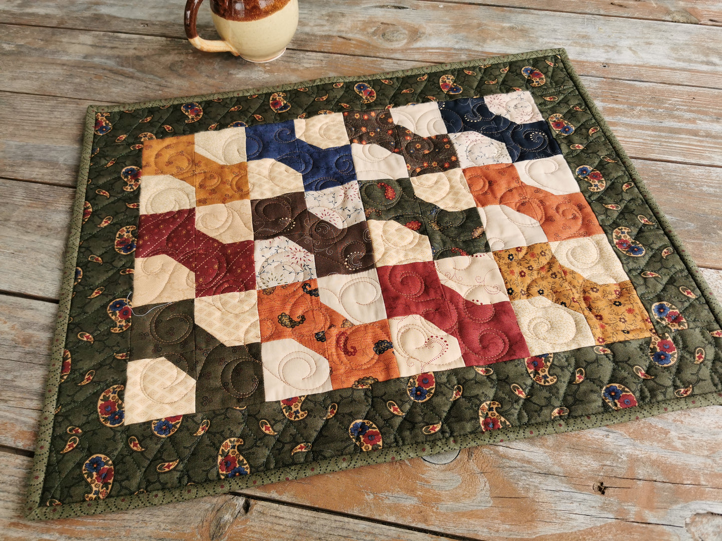 This quilted table runner has an outer border in a forest green paisley print. The patchwork bowties in the center are each a different dark tone colour, and come from a fabric collection called Paisley Park by Kansas Troubles. . Colours include country red, brown, dark blue, burnt orange, antique gold, deep green. The background is done in a scrappy style using various medium tone beige fabrics. A lovely country quilt.