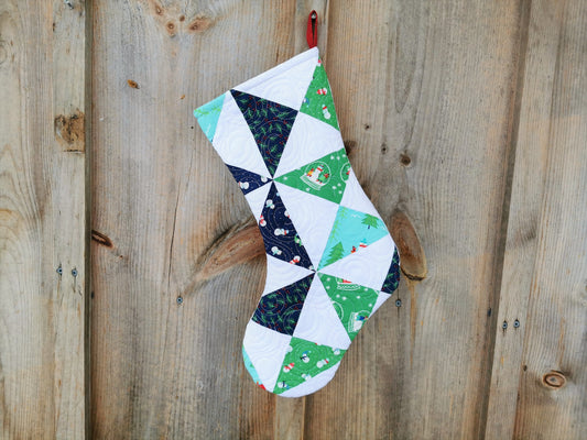 This heirloom quilted stocking has pinwheel piecing in winter themed fabrics on a white background.  Pinwheels are navy, green and a light teal blue. 