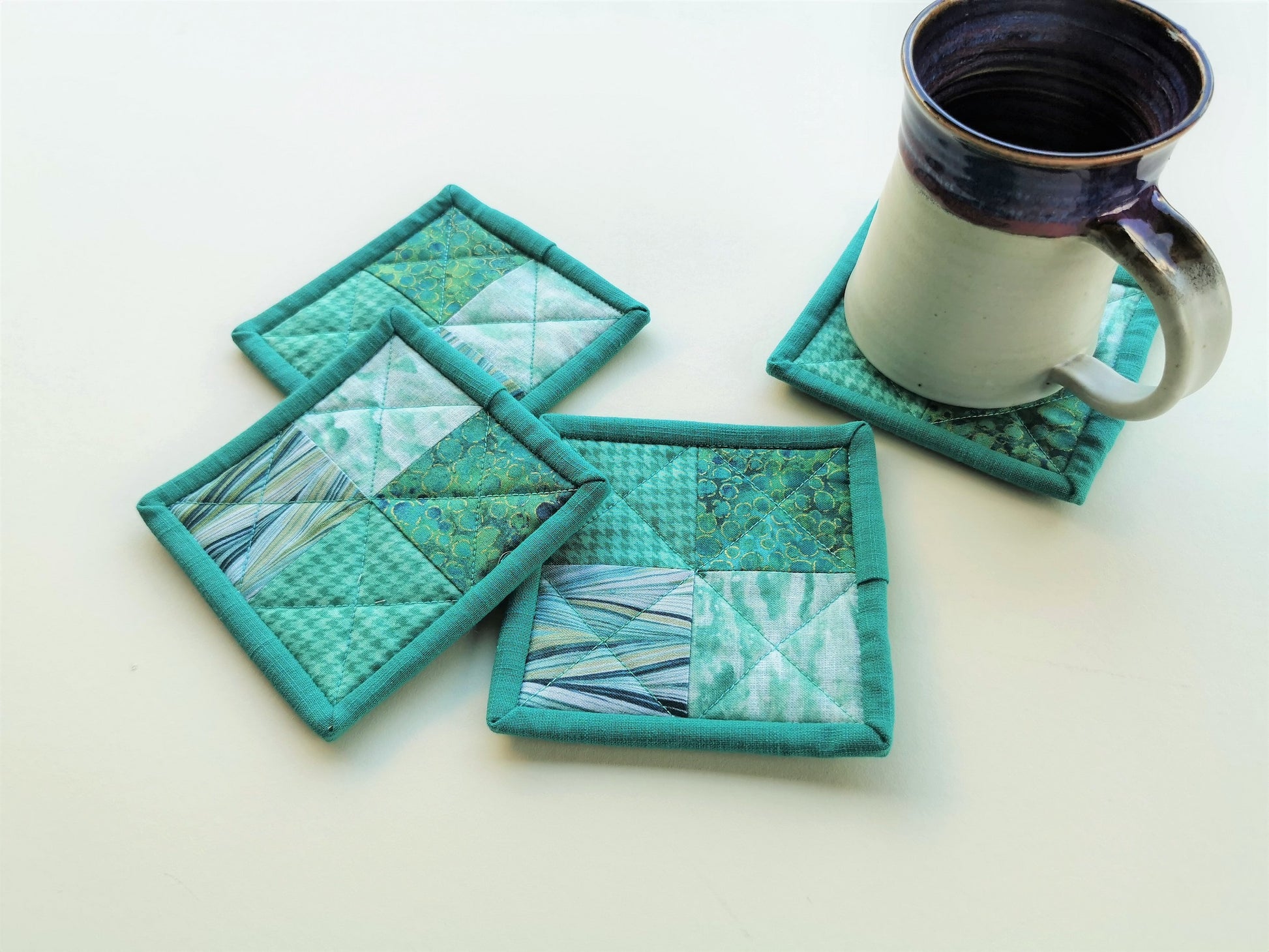 four quilted coasters in modern teal fabrics are displayed with a coffee mug sitting on one of them, showing scale.