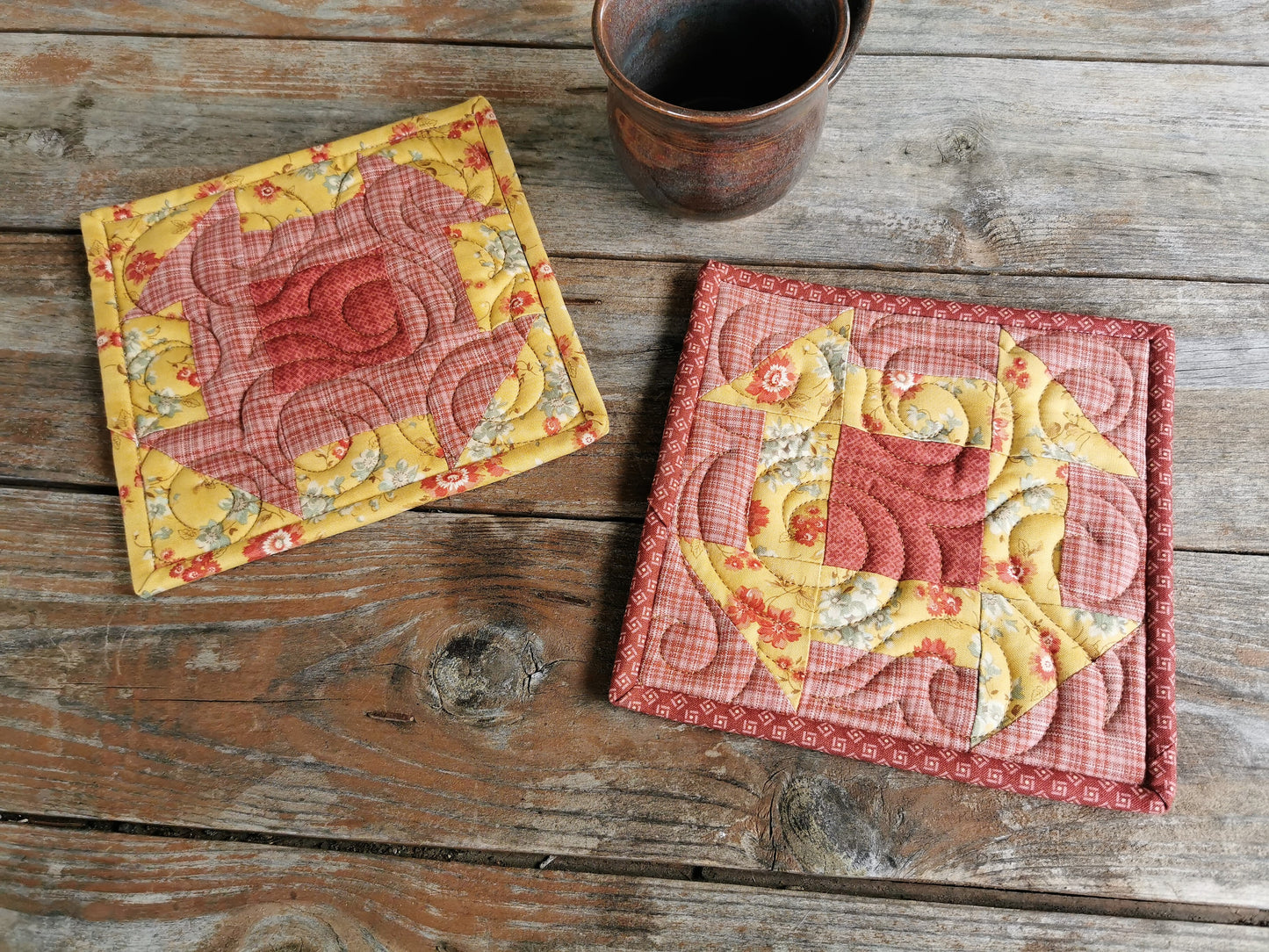 the quilted patchwork potholders are made with fabrics in golden yellow floral and coral pink plaid. They are pieced in the reverse of each other with both having a dark coral pink center block. 