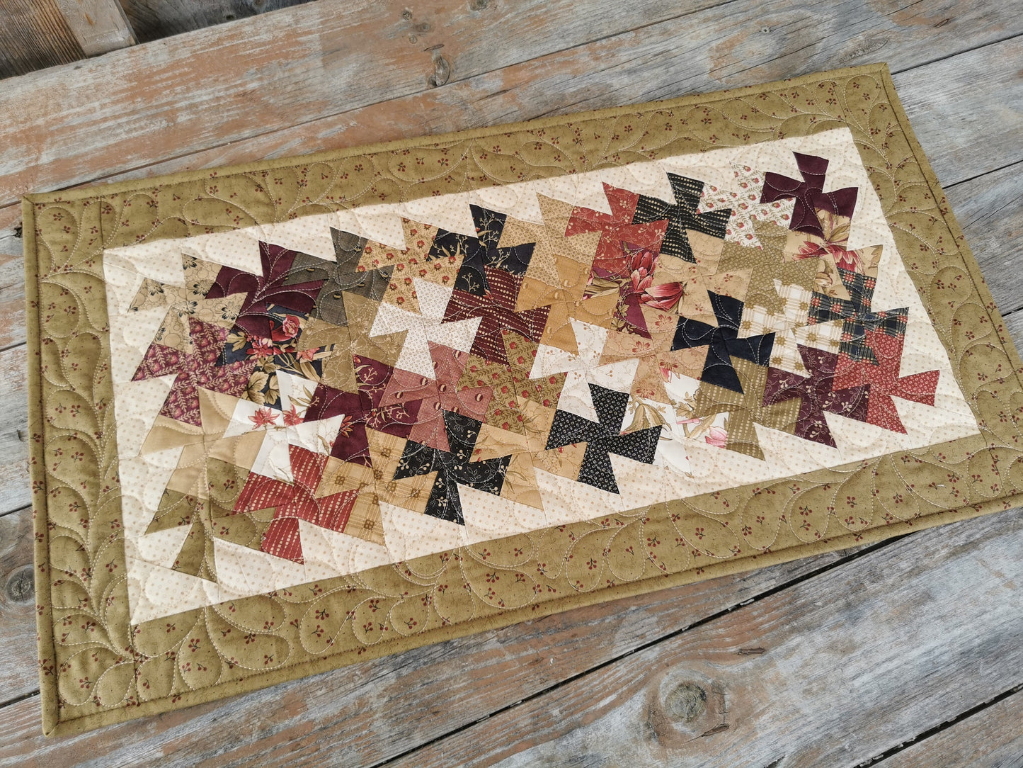The quilted table runner is shown on a wooden table in natural outdoor lighting. The entwining patchwork twisters in country colors are so interesting and pretty. Colors include beige, brown, dark plum, country red, drab khaki green, charcoal. All fabrics are ccoordinating. 