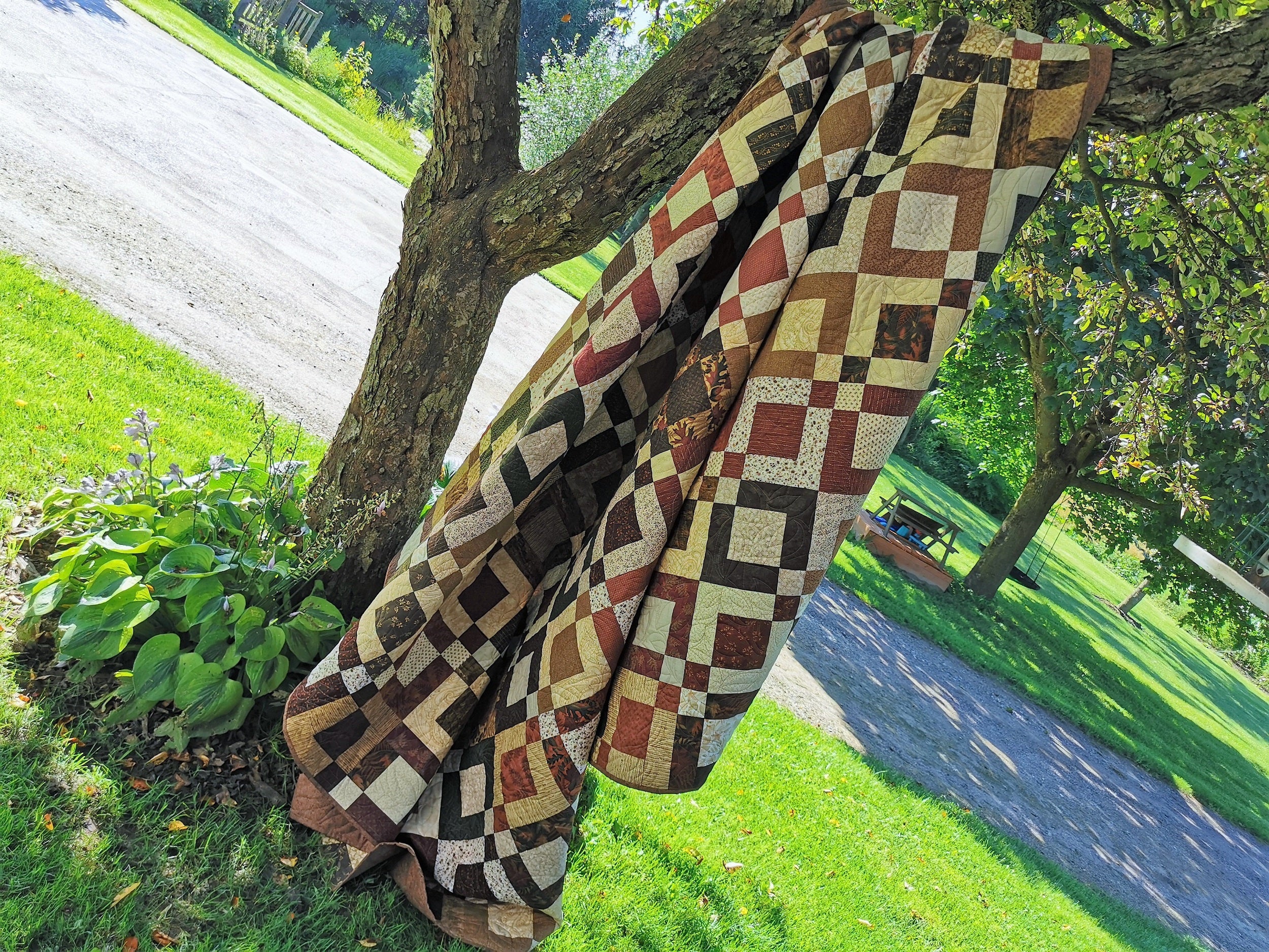 This large queen scrappy patchwork quilt is shown in outdoor light, hanging over a tree branch. Rustic browns, greens, country red, burnt orange, gold and ecru are used in the square patchwork blocks. Pattern looks like squares inside of squares. 