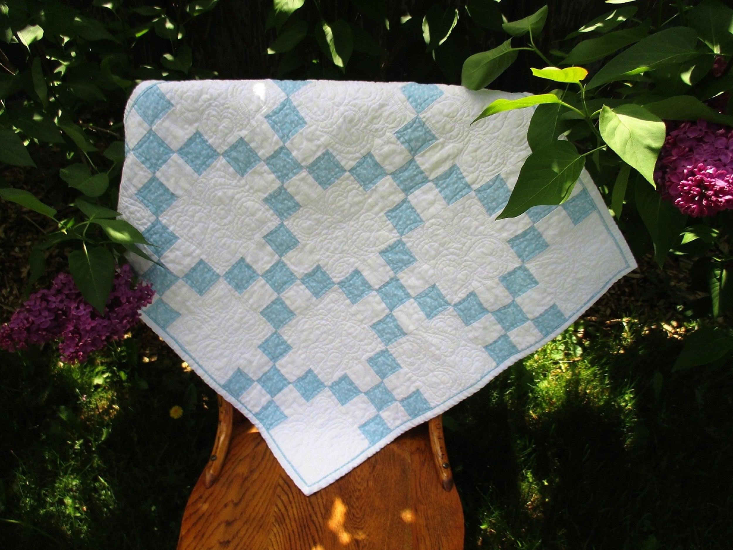 This small blue and white nine patch quilt is shown outdoors over the back of a wooden chair, in front of a lilac bush. 