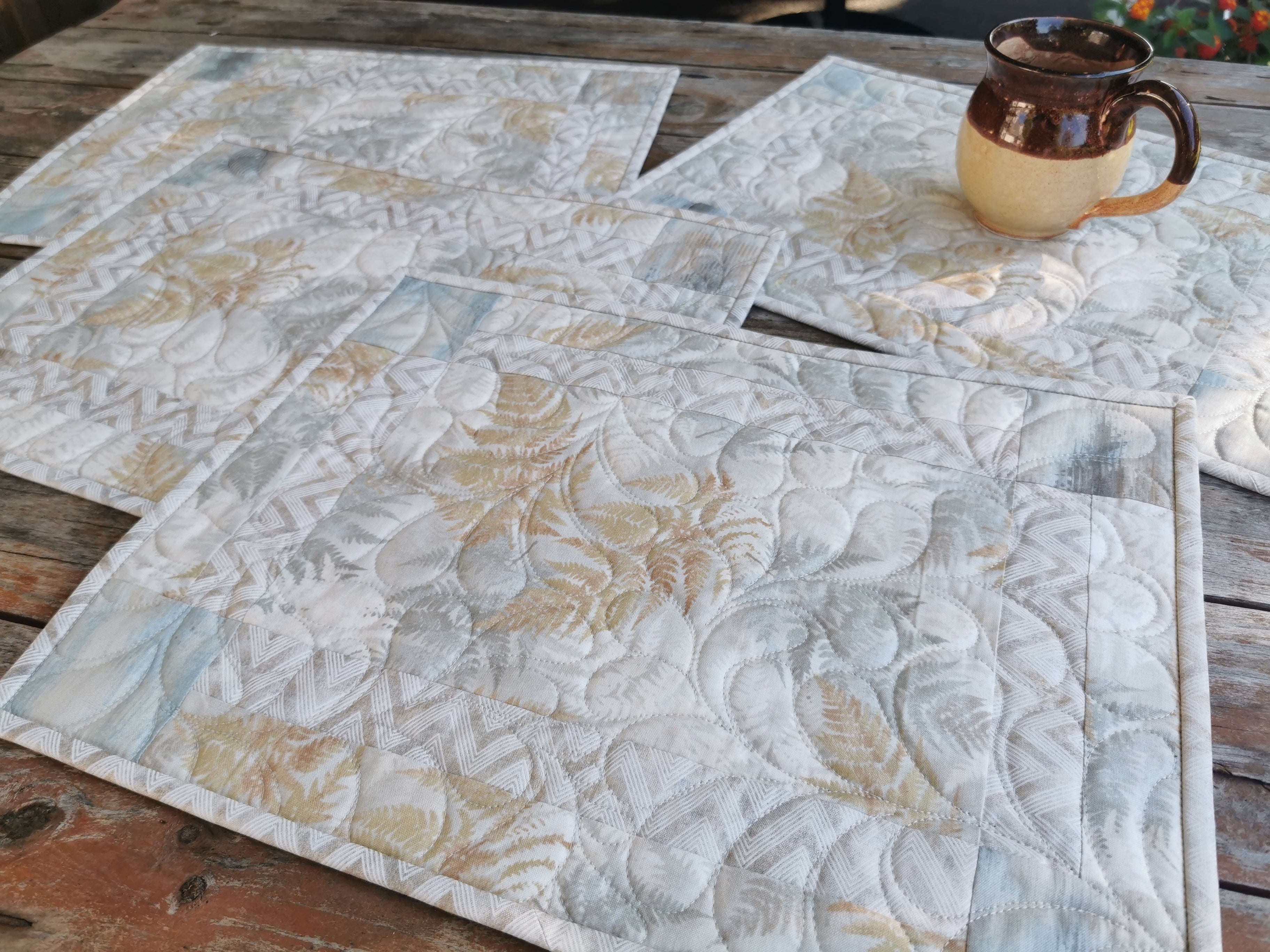 Side angle view of the four quilted placemats. The muted abstract fern pattern features neutral beige and slate blue.