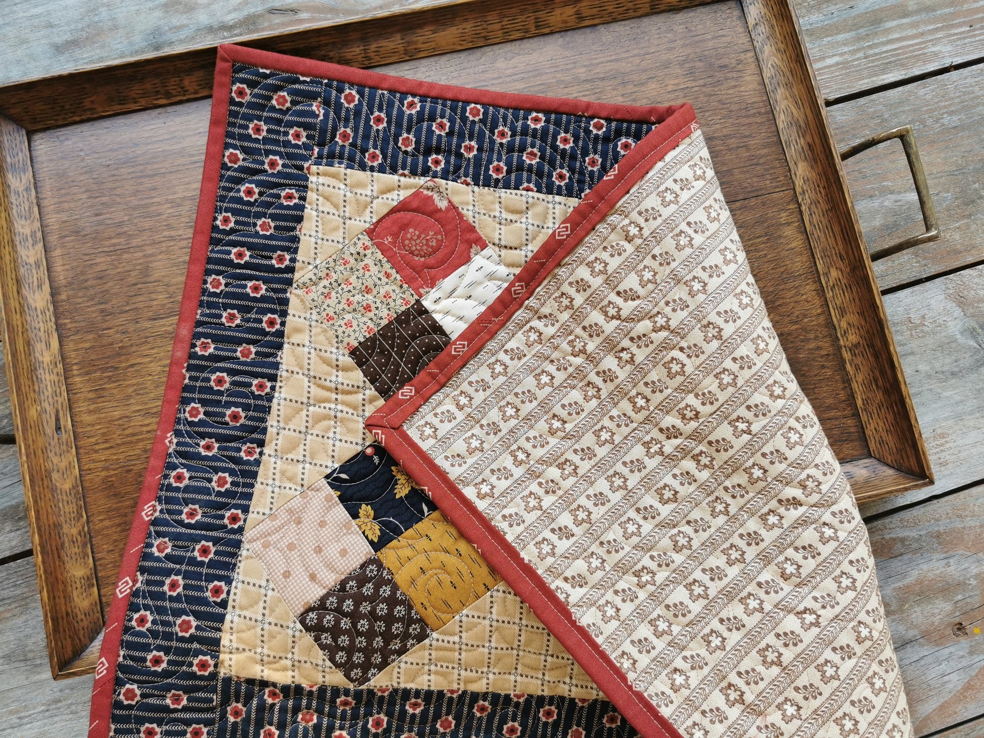 The small scrap quilt is pictured on a wooden tray and folded so that the backing us showing. Backing is a beige print with leafy stripes. All fabrics are coordinating.
