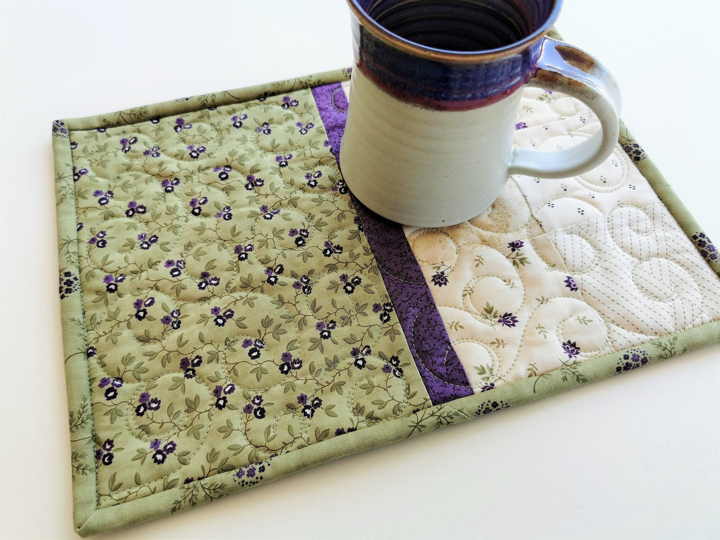the quilted mug rug is shown with a pottery mug sitting on it, showing scale. Quality cotton fabrics are lovely spring green with purple flowers.