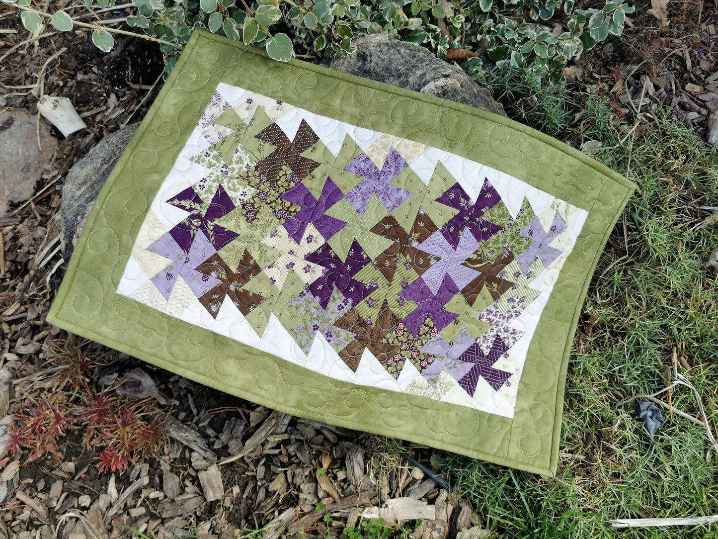 The table runner is shown outdoors, draped over a garden rock. the natural light shows the pretty spring green with purple, brown and cream accents in the scrappy twister piecing in the center of the runner.