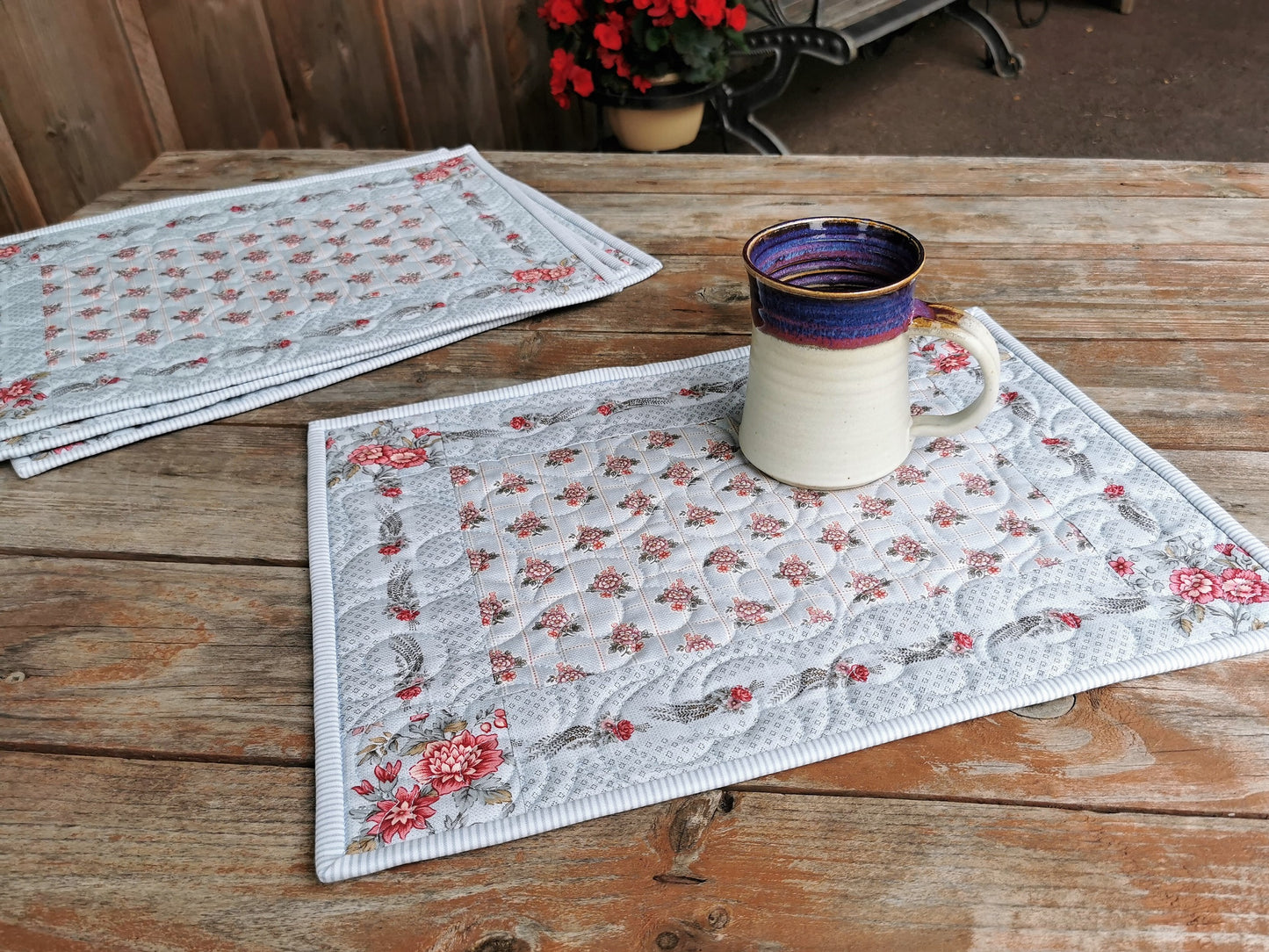 ONe quilted placemat is shown in the foreground with a pottery mug to show scale. Other table mats are stacked in background. This is an outdoor porch setting. 