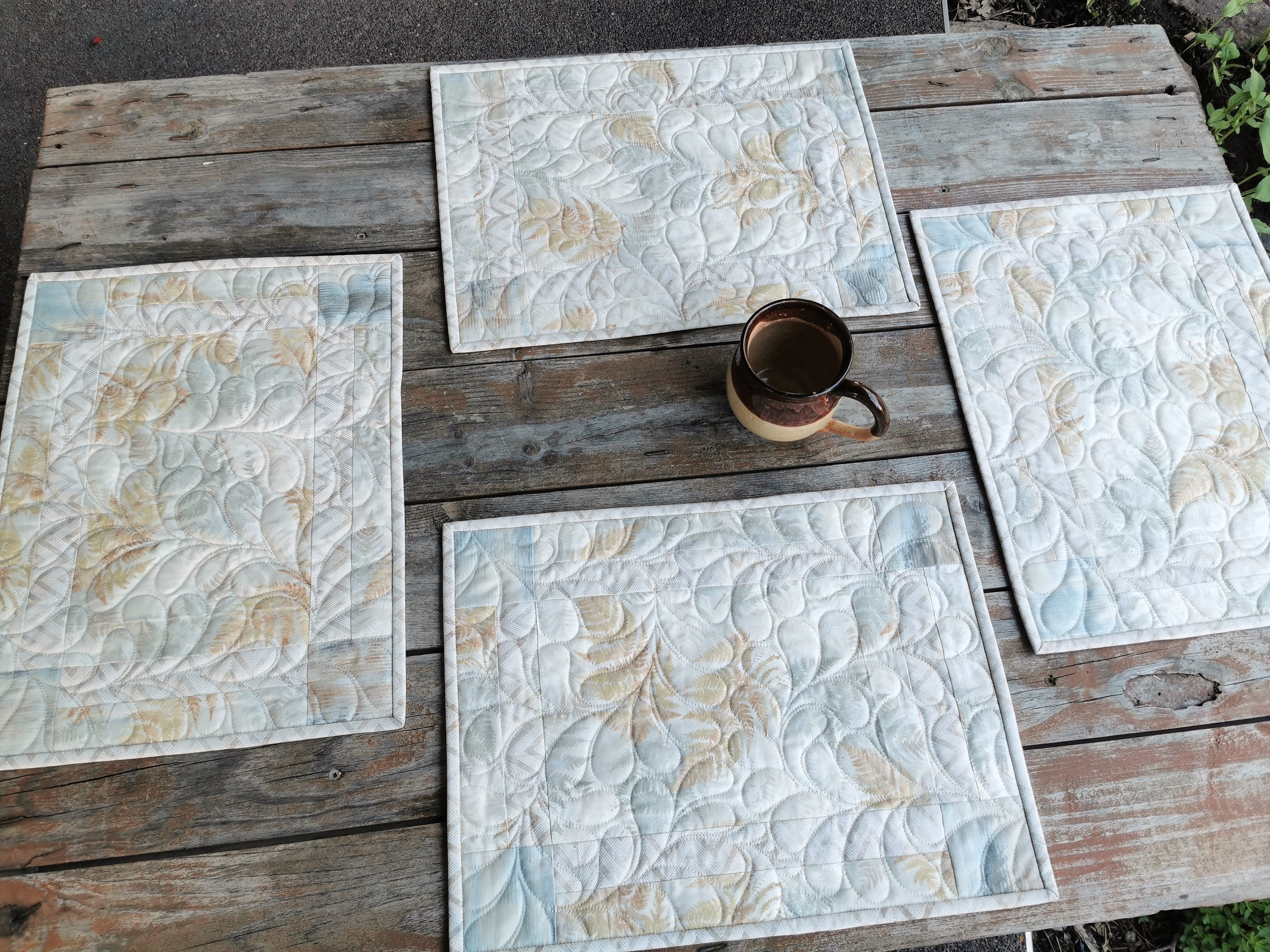 The four quilted placemats are shown on a small table , laid out individually. Feather quilting is visable showing texture.