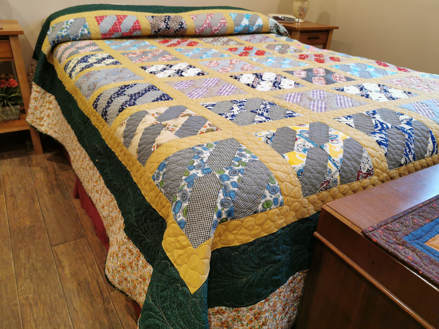 This large queen vintage bed quilt it shown on a queen bed. This view shows along the bedside and how the quitl drapes most of the way to the floor, and it tucked up and over the pillows. The middle (top of the bed)  quilt top has the scrappy vintage patchwork blocks set in a mustard yellow grid pattern. This grid of scrappy blocks is surrounded by a dark green border then a beige floral border around the outside