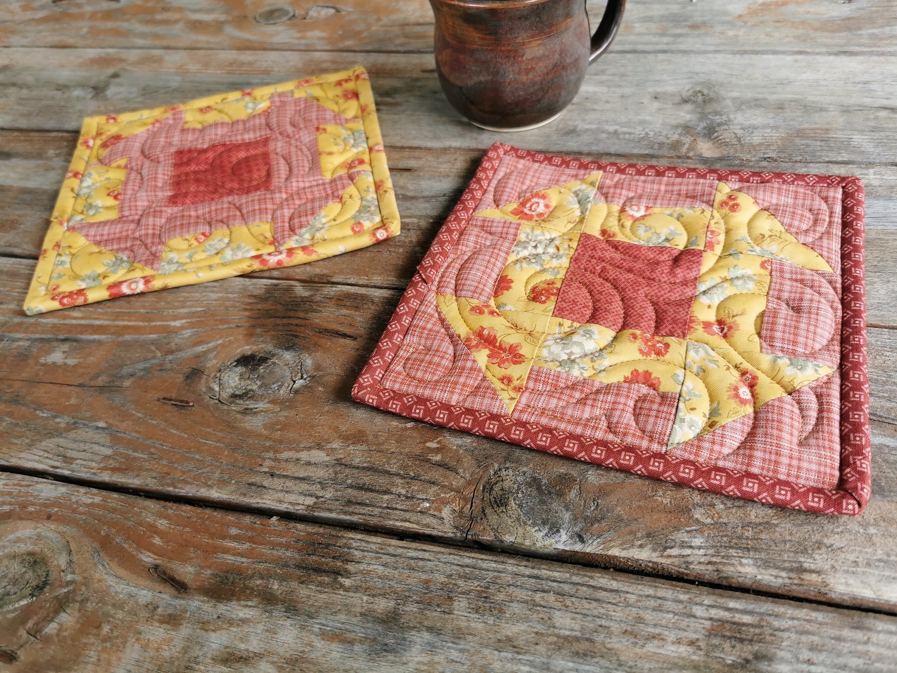 the quilted patchwork potholders are made with fabrics in golden yellow floral and coral pink plaid. They are pieced in the reverse of each other with both having a dark coral pink center block. This view is shown looking at the potholders from a side angle