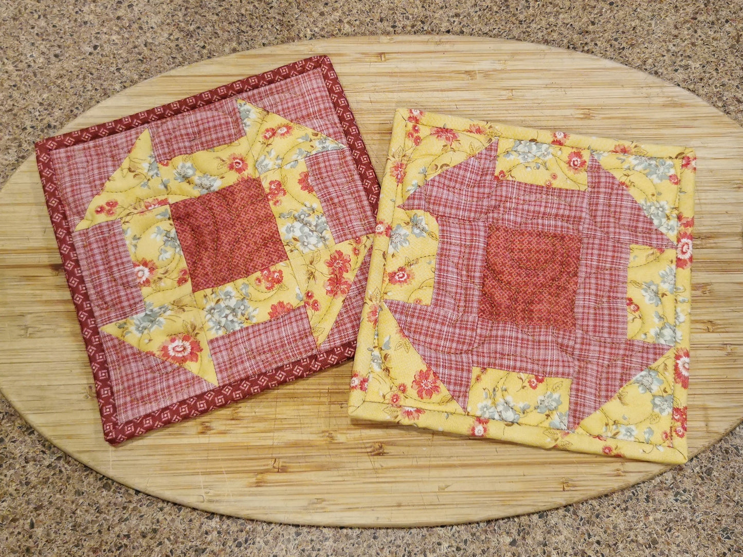 the quilted patchwork potholders are made with fabrics in golden yellow floral and coral pink plaid. They are pieced in the reverse of each other with both having a dark coral pink center block. This view is shown in indoor lighting with the quilting texture minimized so that the patchwork is prominent. 