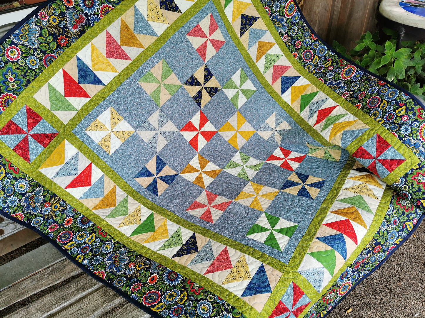 This bright pinwheel quilt is framed by a beautiful blue floral print. The border colors are repeated throughout the the triangle piecing in the main part of the quilt.  Happy primary colors of red, green, yellow, blue, pink, gold. Main color is blue with various shades of the other  colors. 
