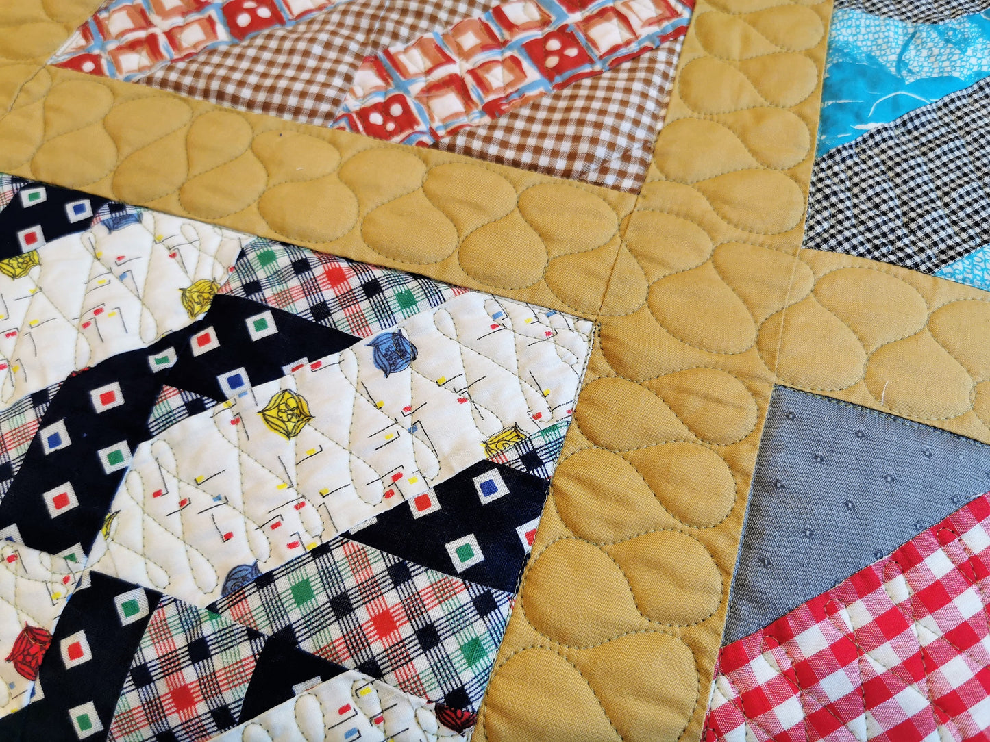this is a close up of a small portion of the vintage bed quilt top showing the mustard yellow grid with the serpentine stitching on it. A few of the vintage fabrics that make up the scrappy blocks, that the quilt is built around, are also shown.  there is red gingham, brown gingham, black gingham, gray, navy print, red and blue print, navy  plaid, bright blue. 