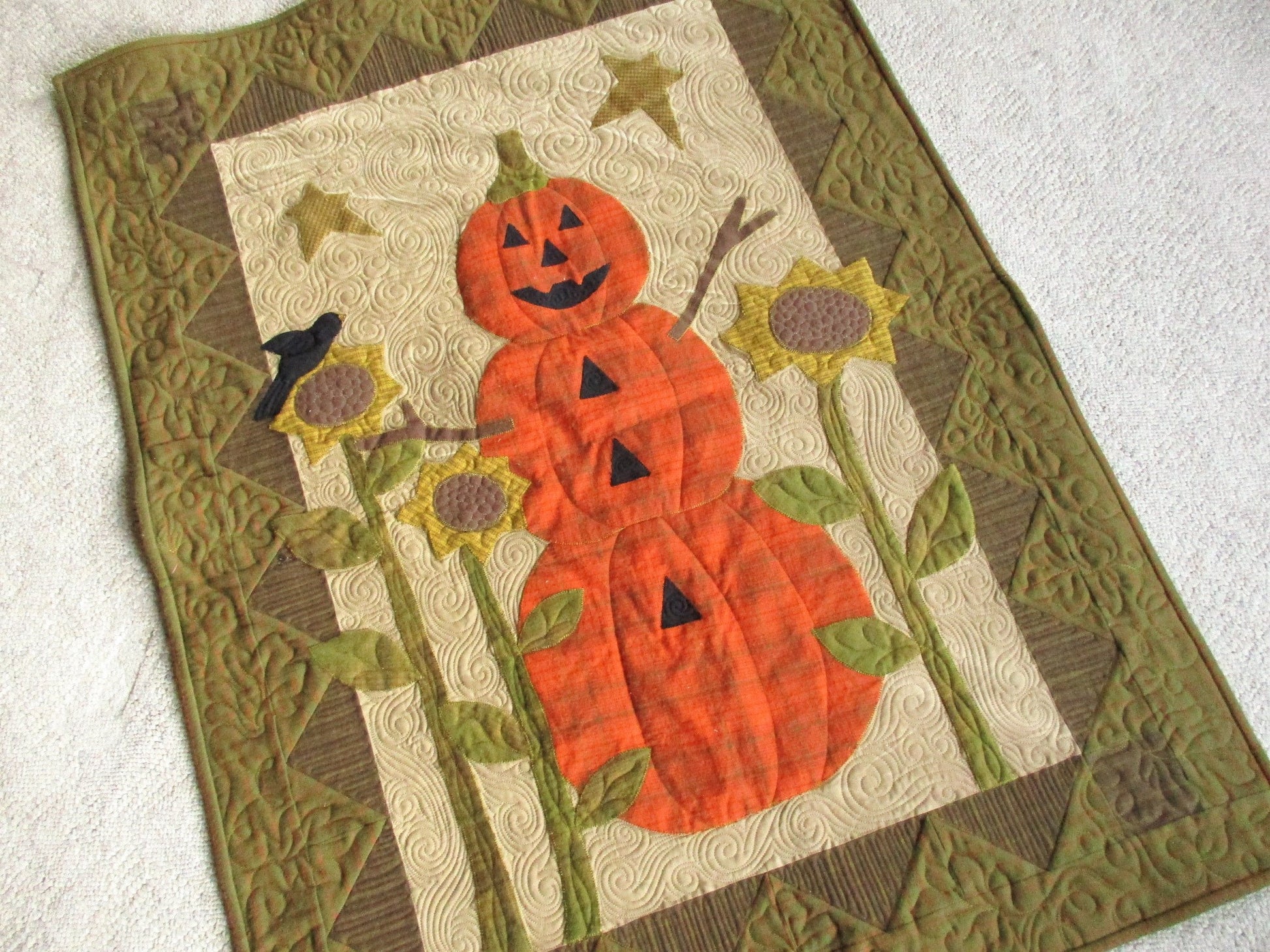 A large rustic quilted wall hanging features appliqued design elements. Find a pumpkin man with a jack-o-lantern face, tall sunflowers on stems with a crow sitting on one and stars in background. Beautiful custom quilting is a key feature of this wall quilt. Colors include burnt orange for the pumpkin man, antique gold sunflowers, drab olive green and brown and a tan background. 