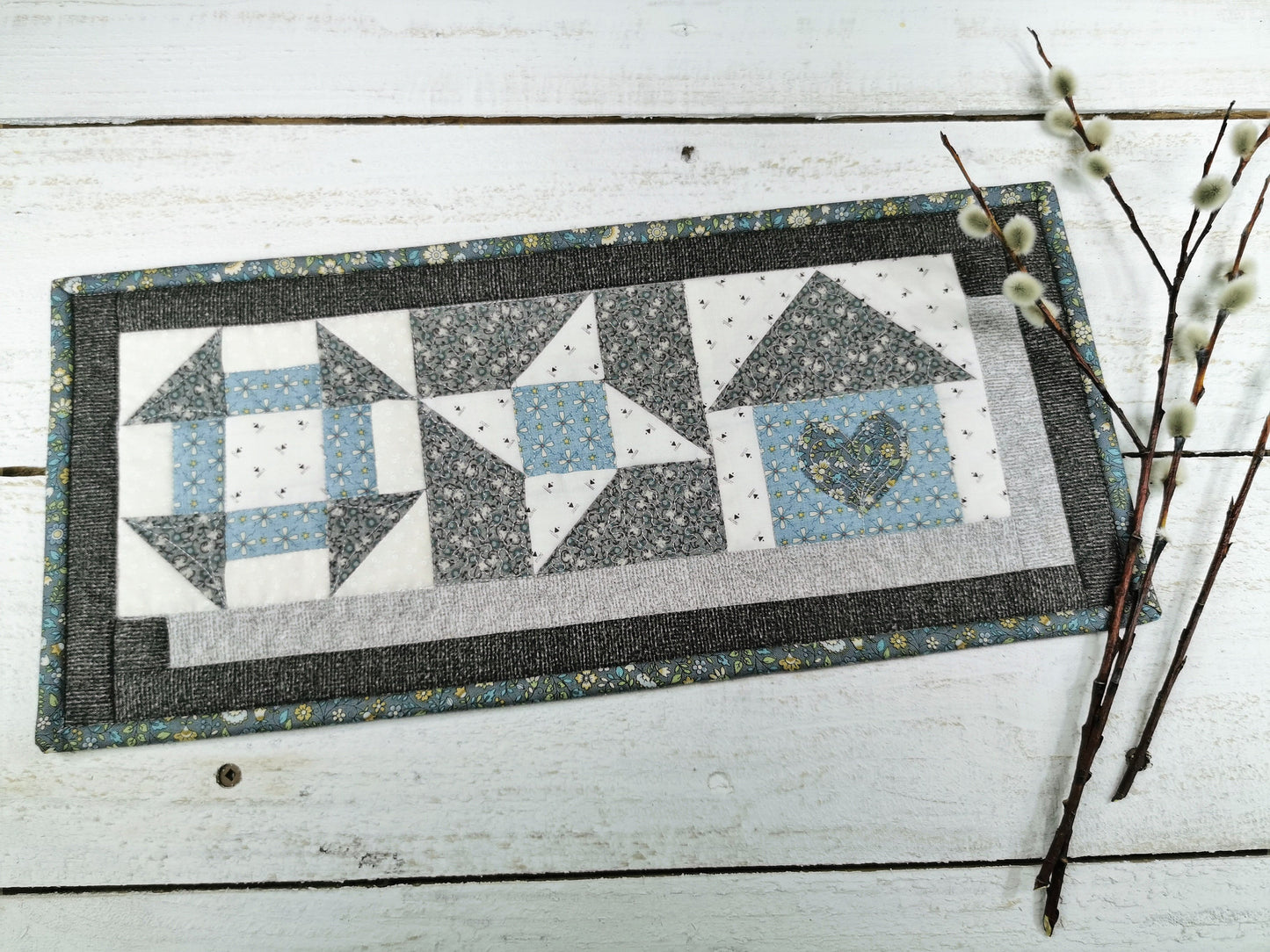 An overhead view of the patchwork mini quilt with pussywillow branches laying along the right of photo. A pretty gray and blue mini sampler style is made with three different quilt blocks in a horizontal row. The blocks, left to right, are churn and dash, friendship star and a house block. 