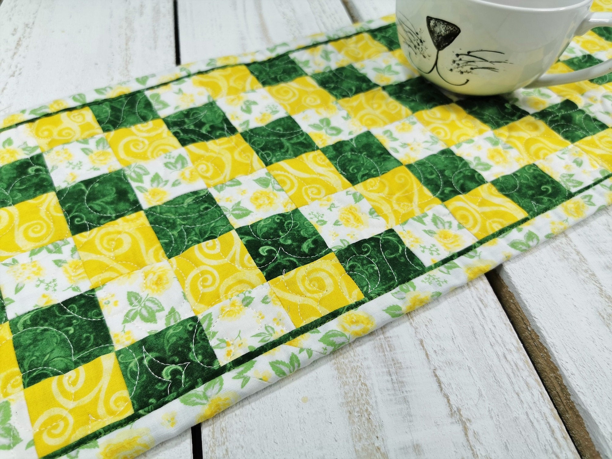 quilted table runner shown on white wooden table with a cat face mug in upper right corner showing scale.