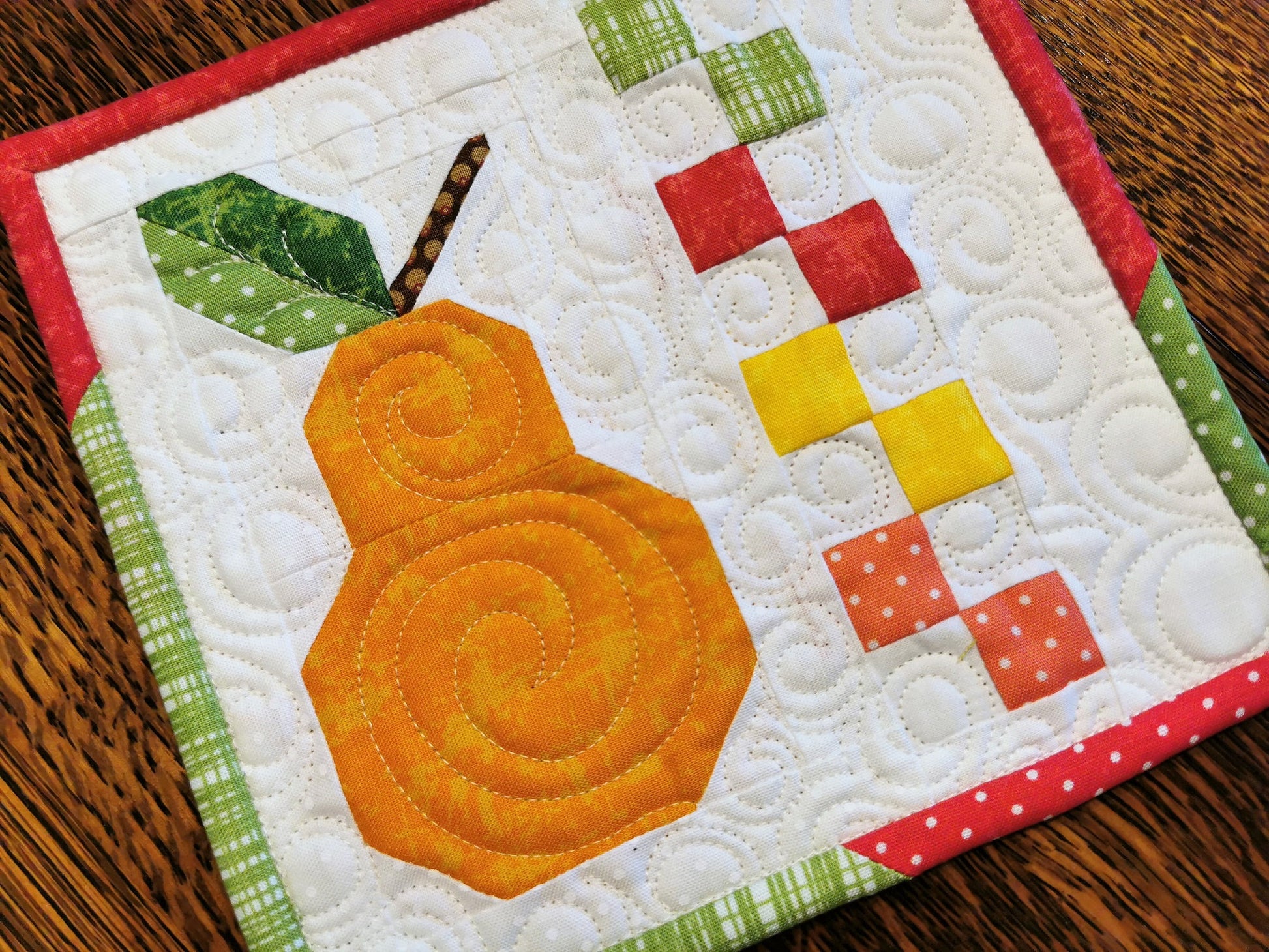 This bright orange patchwork pear is made of small, intricate piecing. Bright primary colors make up the rest of the patchwork piecing on a white background filled with custom quilting. Colors include red, green, yellow, coral pink.