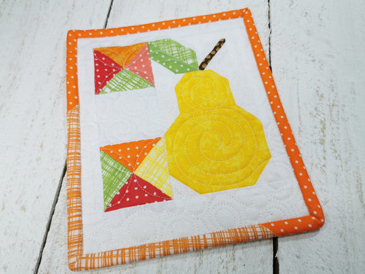 Overhead view of this small patchwork pear quilt. Perfect for a mug rug the primary colors are cheerful and fun. Bright yellow, orange, red and green on a white background are eye catching. Beautiful quilting adds detail and texture..