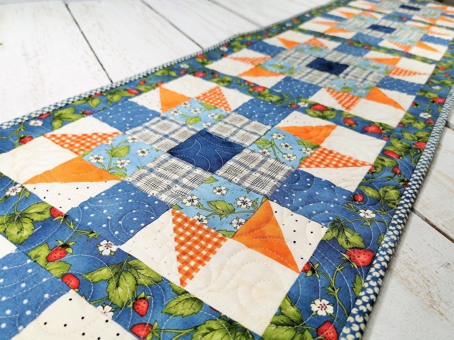 This long table runner features a strawberry focal fabric. A variety of pretty blue fabrics are complimented by orange and cream creating a lovely, summery table runner. 